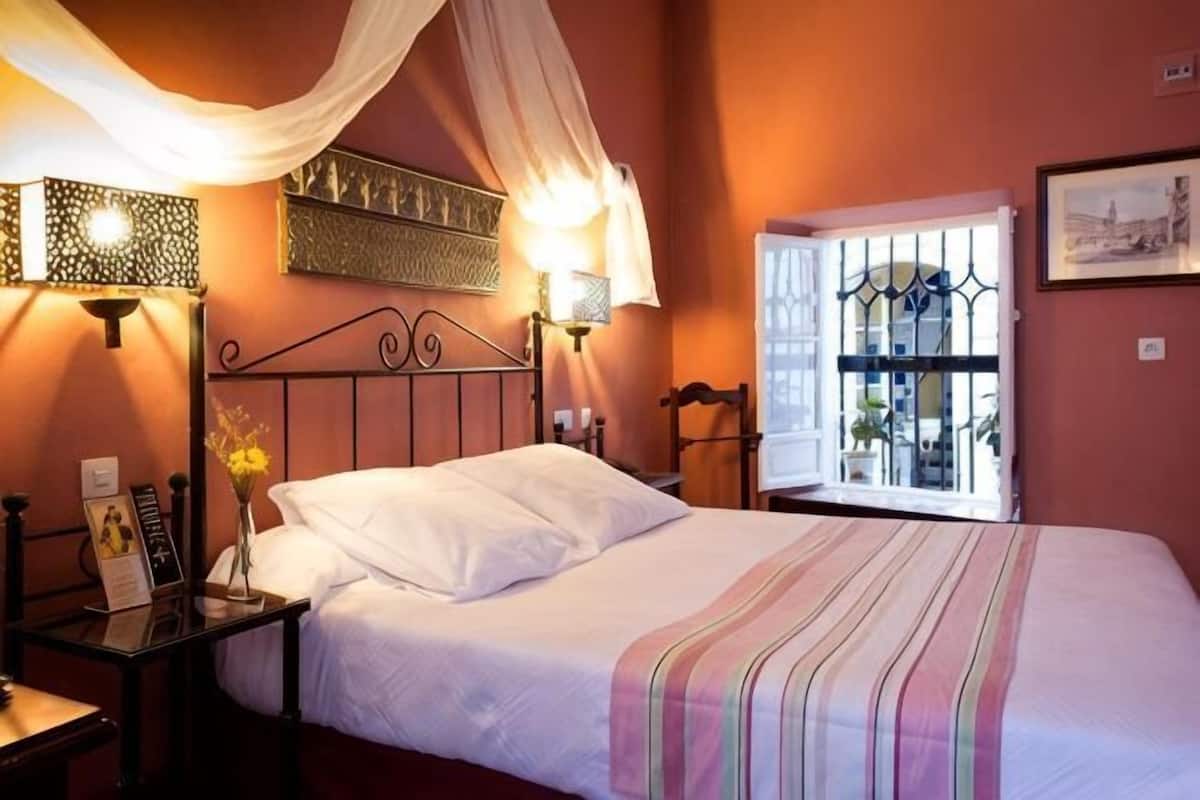 Un dormitorio con cama, lámparas de noche, mesita de noche, ventana con rejas y cortina.