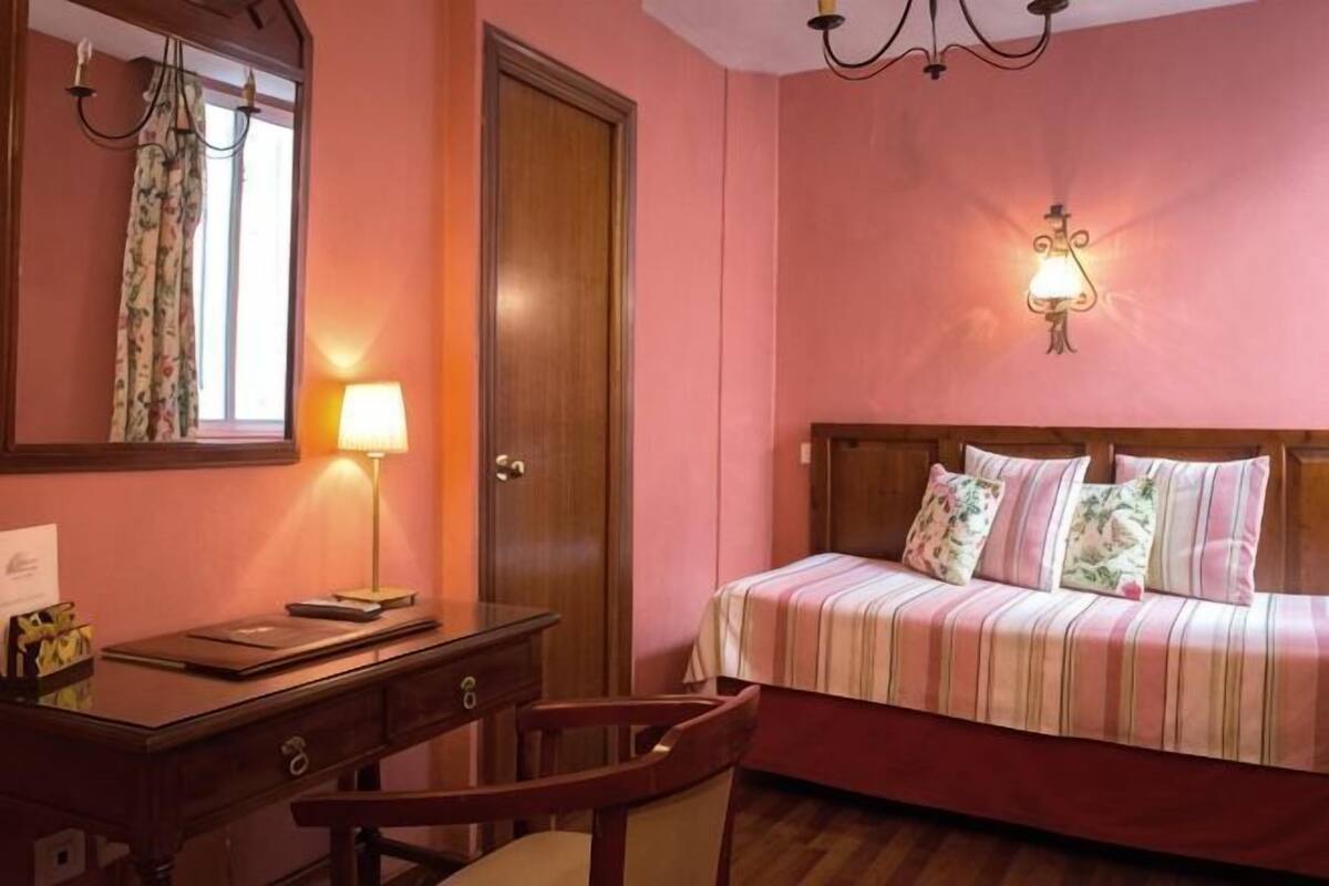 Un dormitorio con cama, escritorio, silla, espejo y lámpara de pared.