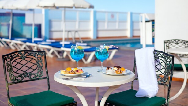 Une table ronde avec deux assiettes de nourriture et deux boissons bleues, entourée de chaises vertes à coussins et de parasols blancs.