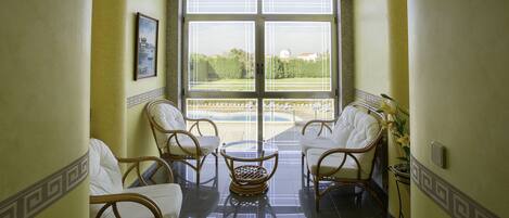 Een balkon met rieten stoelen en een tafel, een ingelijst kunstwerk aan de muur, en uitzicht op groen en gebouwen.
