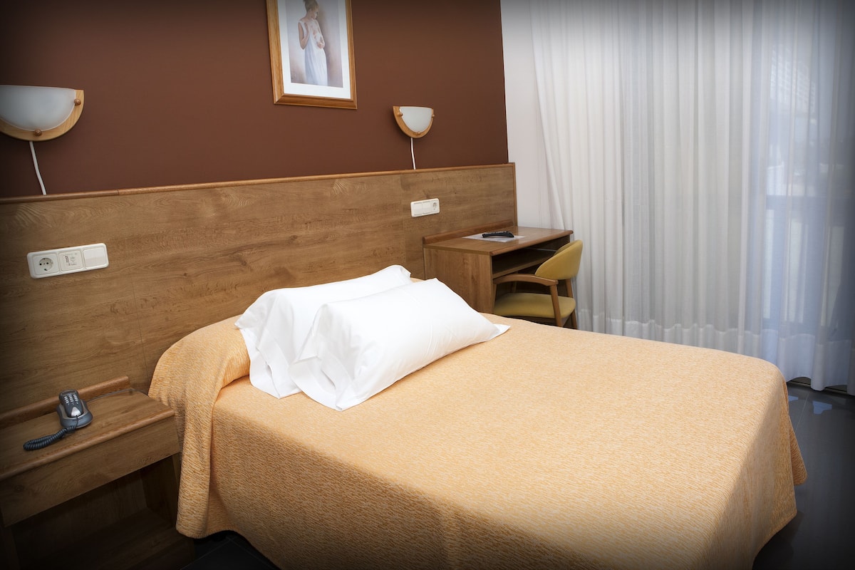 Een hotelkamer met een houten hoofdeinde, een bed met een witte dekbedovertrek, een nachtkastje met een lamp en een bureau met een stoel.