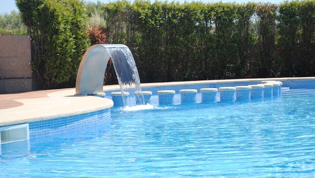 Una piscina con un elemento acuático, rodeada por una solana con baldosas y un seto.