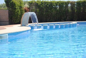 Une piscine agrémentée d’un élément décoratif aquatique, entourée d’une terrasse carrelée et d’une haie.