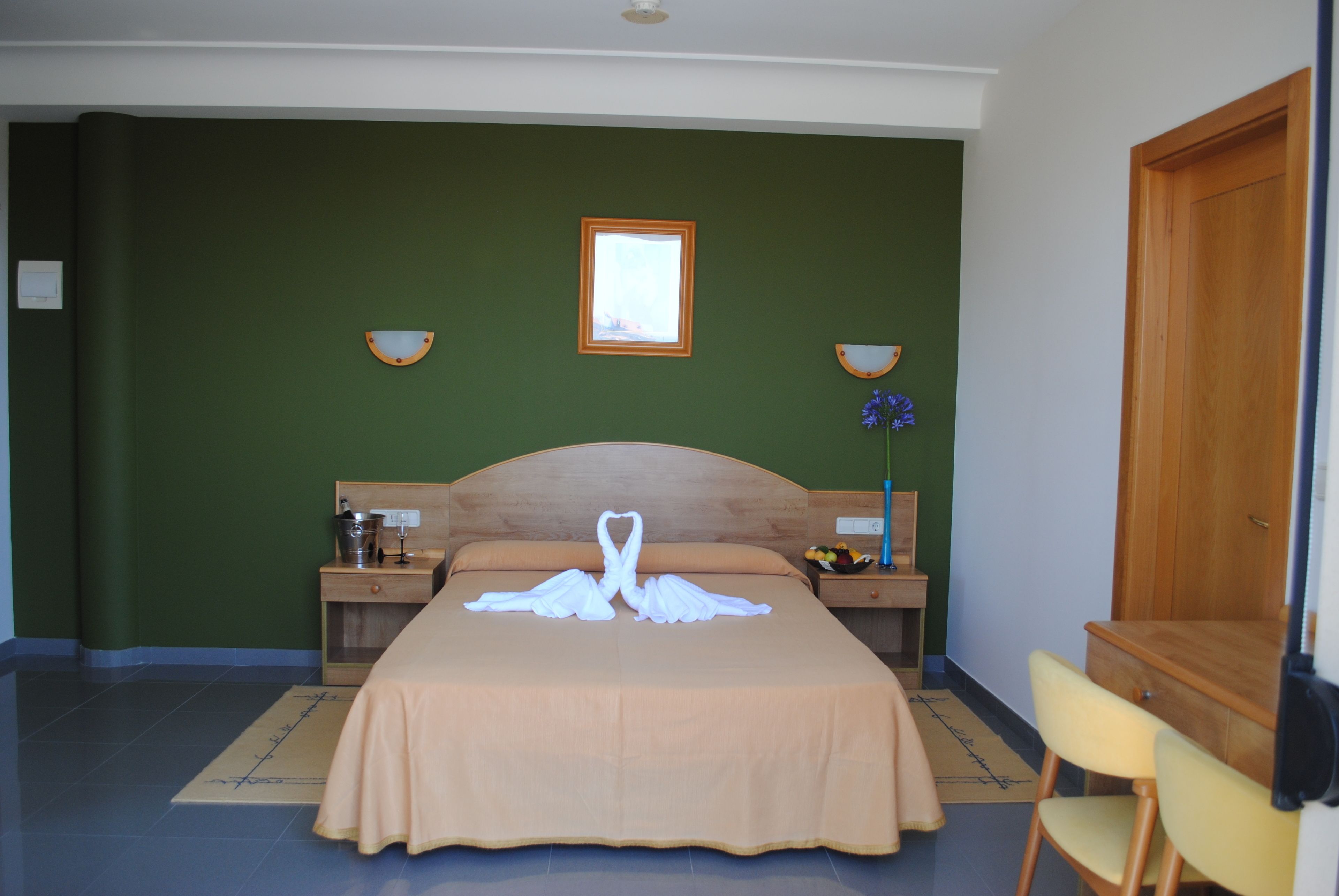 Habitación de hotel con una cama, cabecero de madera, dos mesitas de noche con lámparas, una mesita auxiliar de madera, una silla y una puerta de madera.
