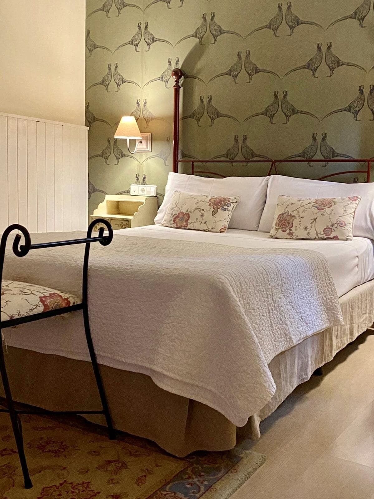 Una cama impecablemente hecha con almohadones florales, una mesita de noche con lámpara y un papel tapiz estampado con motivos de pájaros.