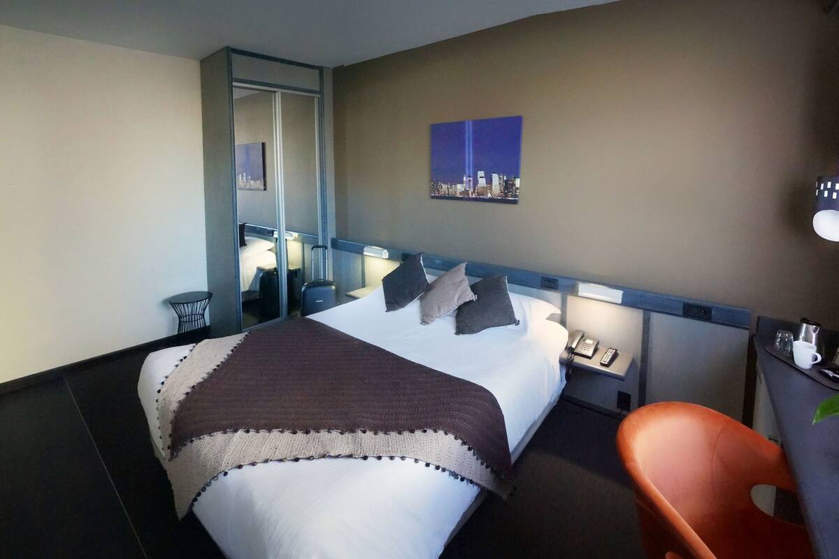 A hotel room with a bed, bedside table, chair, and a cityscape painting on the wall.