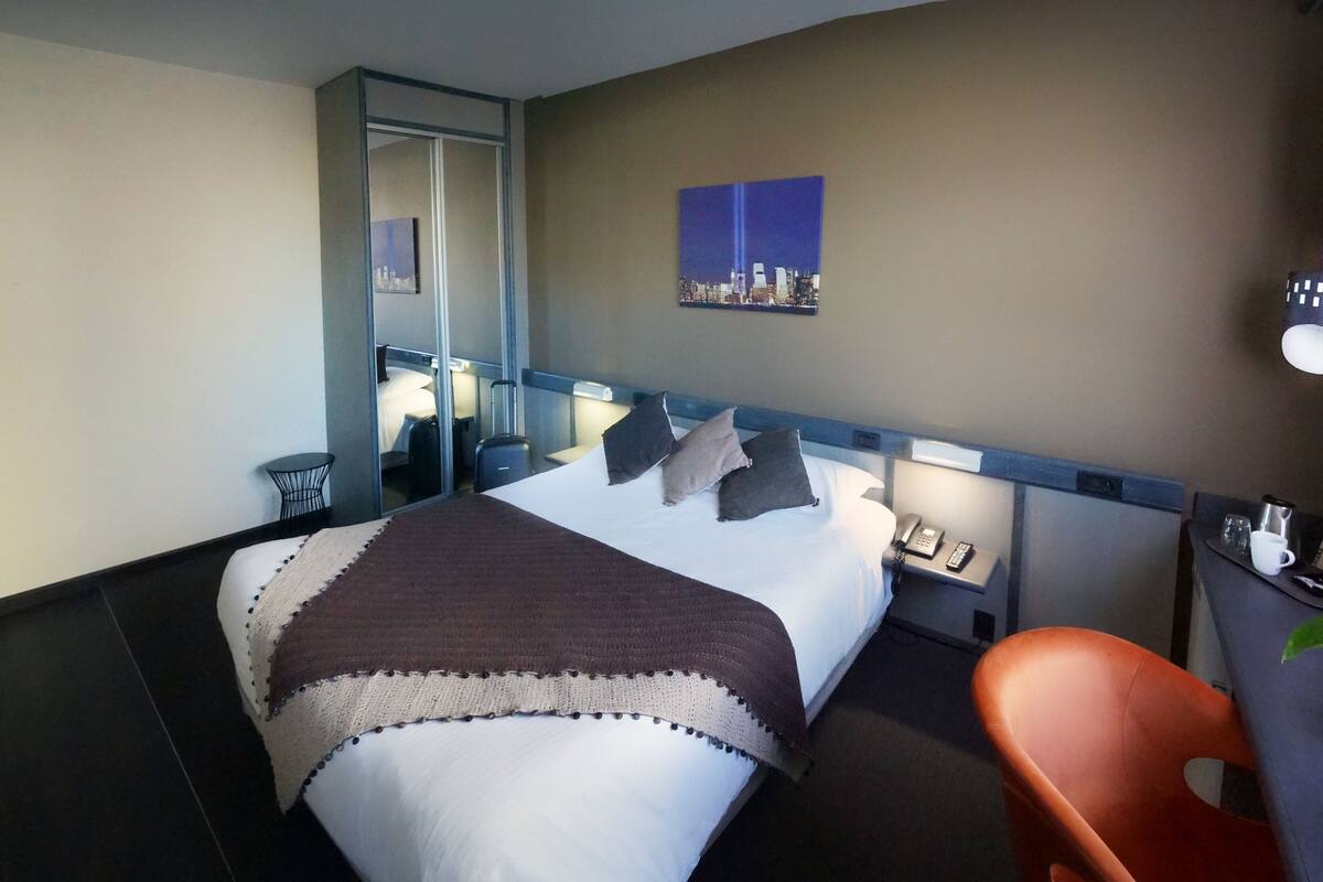 A hotel room with a bed, a chair, a small table, a nightstand, a painting, and a cityscape view.
