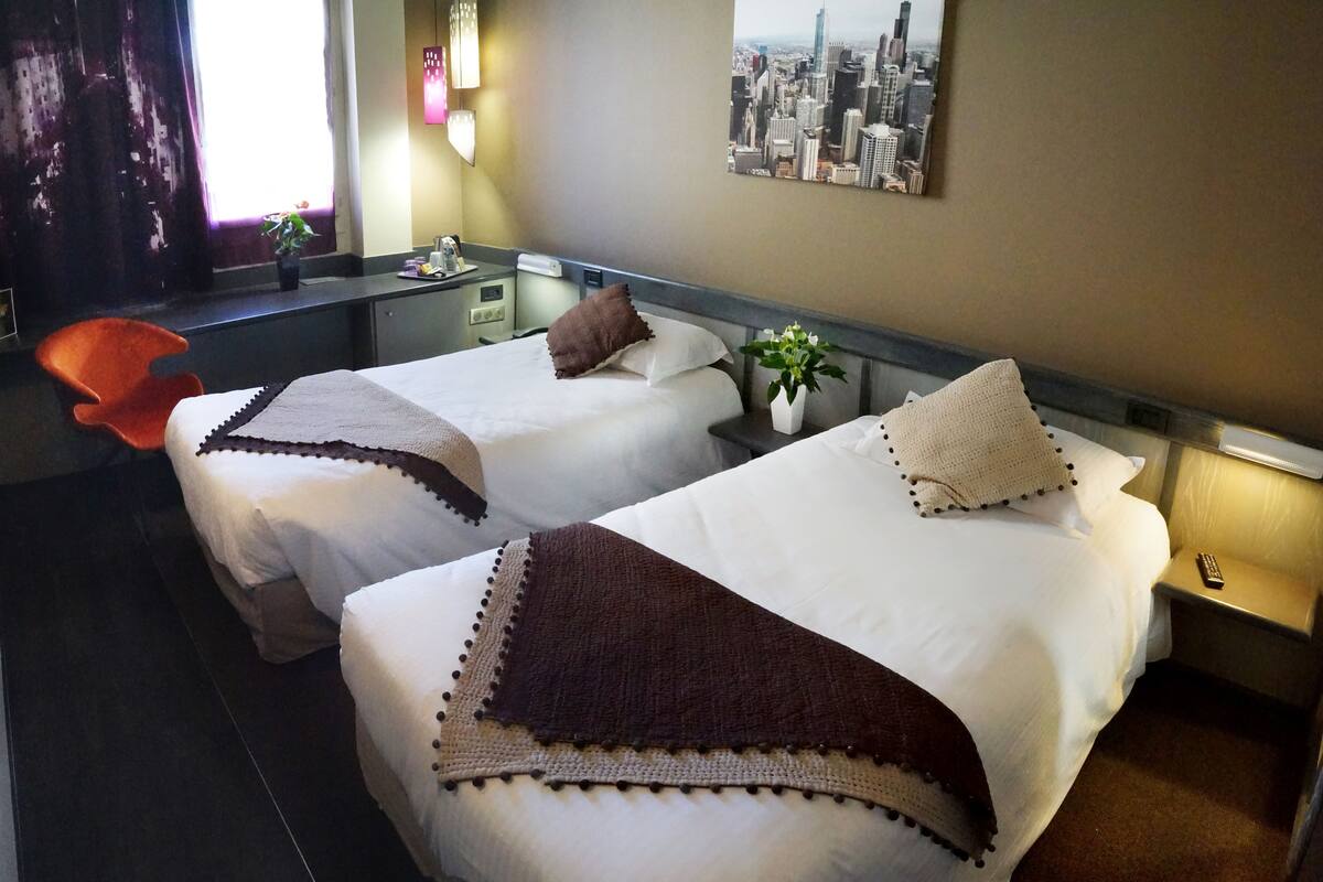 A hotel room with two beds, a desk, a chair, a cityscape painting, and a window with curtains.