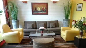 A living room with a sofa set, two armchairs, a coffee table, and a round ottoman. There are two large potted plants, one on each side of the sofa. A framed painting hangs on the wall above the sofa. - Hotel Carmin (Le Havre)