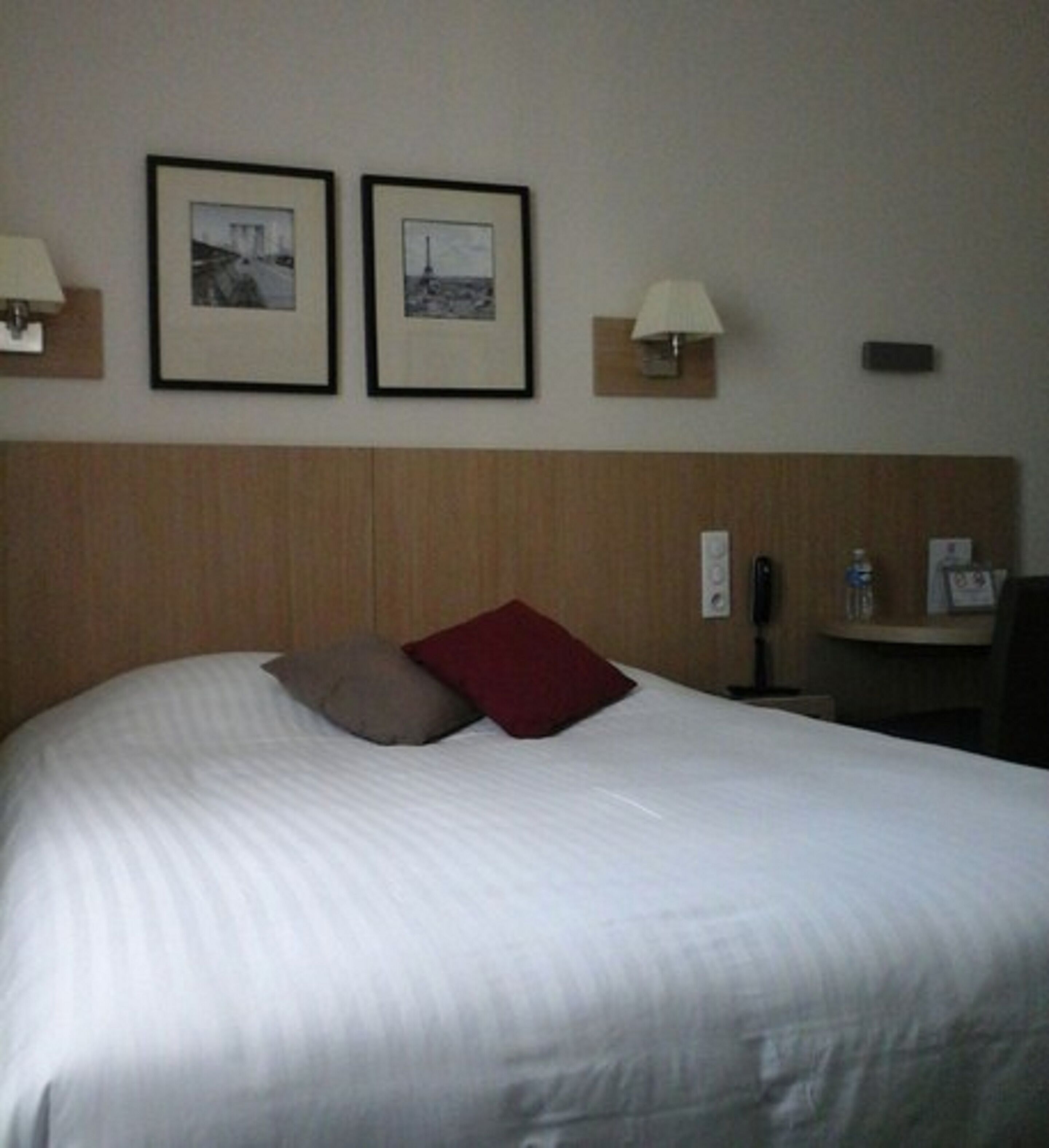 Une chambre d’hôtel avec un grand lit, deux tableaux encadrés au mur, une lampe de chevet et une petite table sur laquelle se trouve une bouteille.