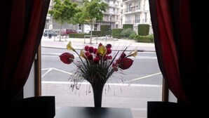 A window with red curtains, a vase of flowers, and a view of a street with buildings and parked cars. - Hotel Carmin (Le Havre)