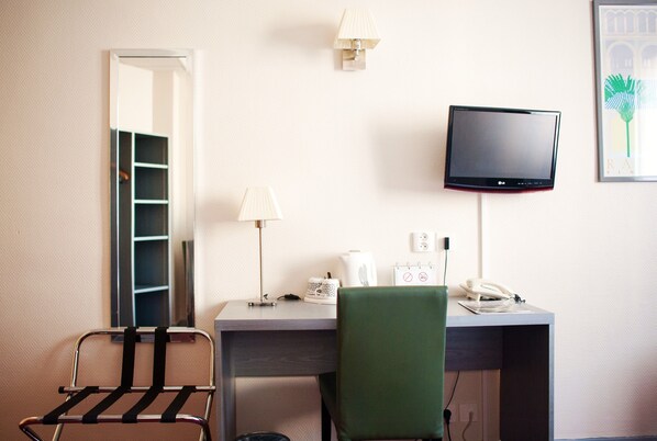 A hotel room with a desk, chair, lamp, phone, and a TV mounted on the wall.