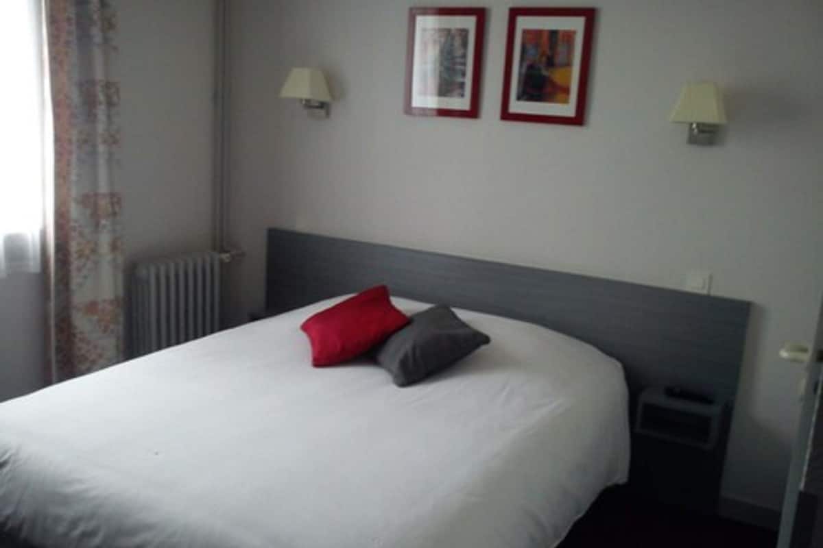 Una cama bien hecha con edredón blanco, un almohábil rojo y un almohábil gris. Hay dos lámparas de pared a cada lado de la cama.