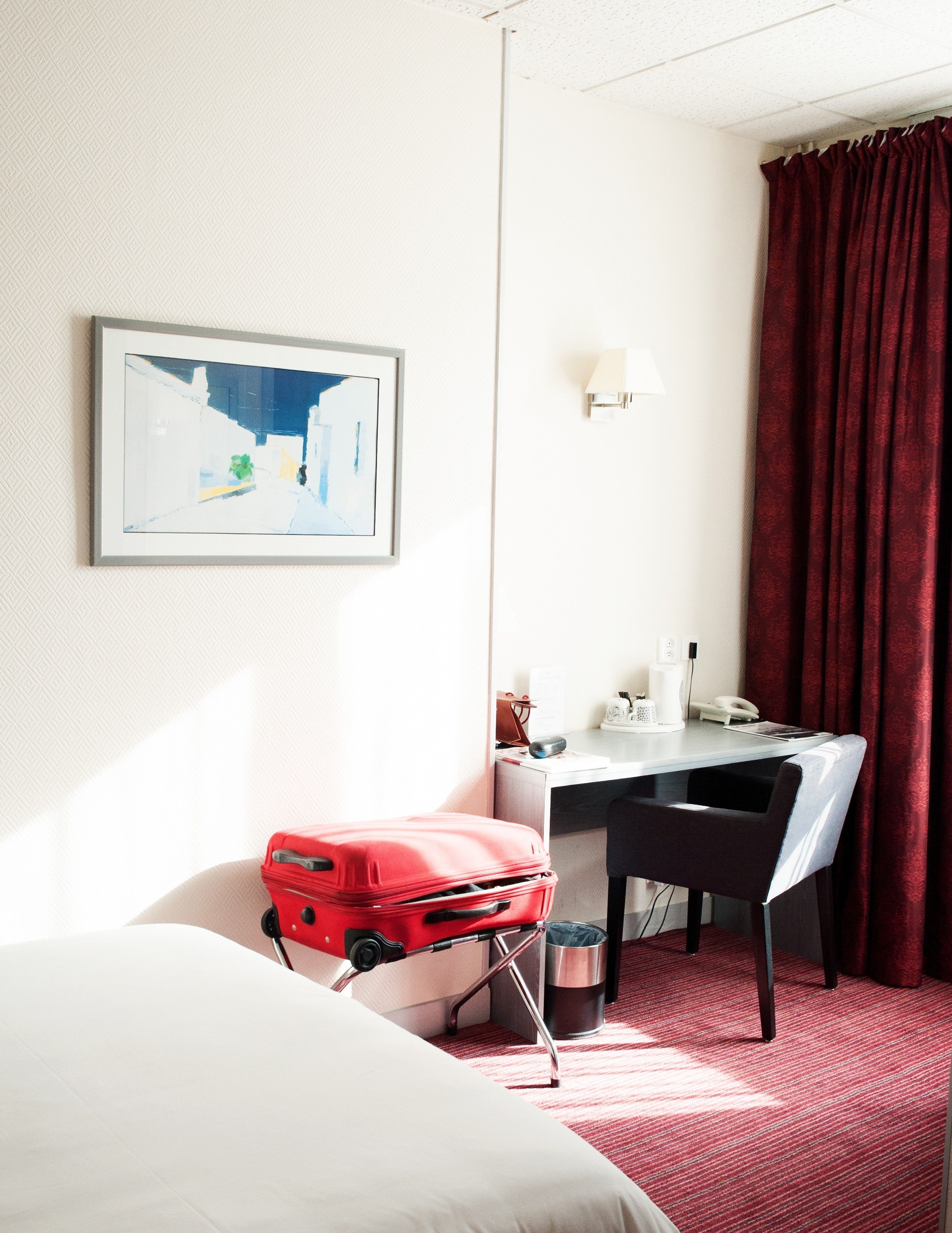 Quarto de hotel com cama, escrivaninha, cadeira, mala vermelha, lixeira e um quadro na parede.