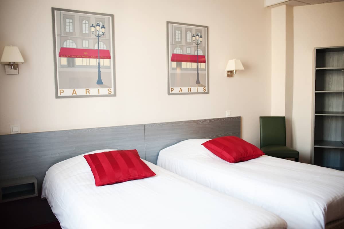 Habitación de hotel con dos camas, cada una con un almohadón rojo, y dos carteles enmarcados de París en la pared.