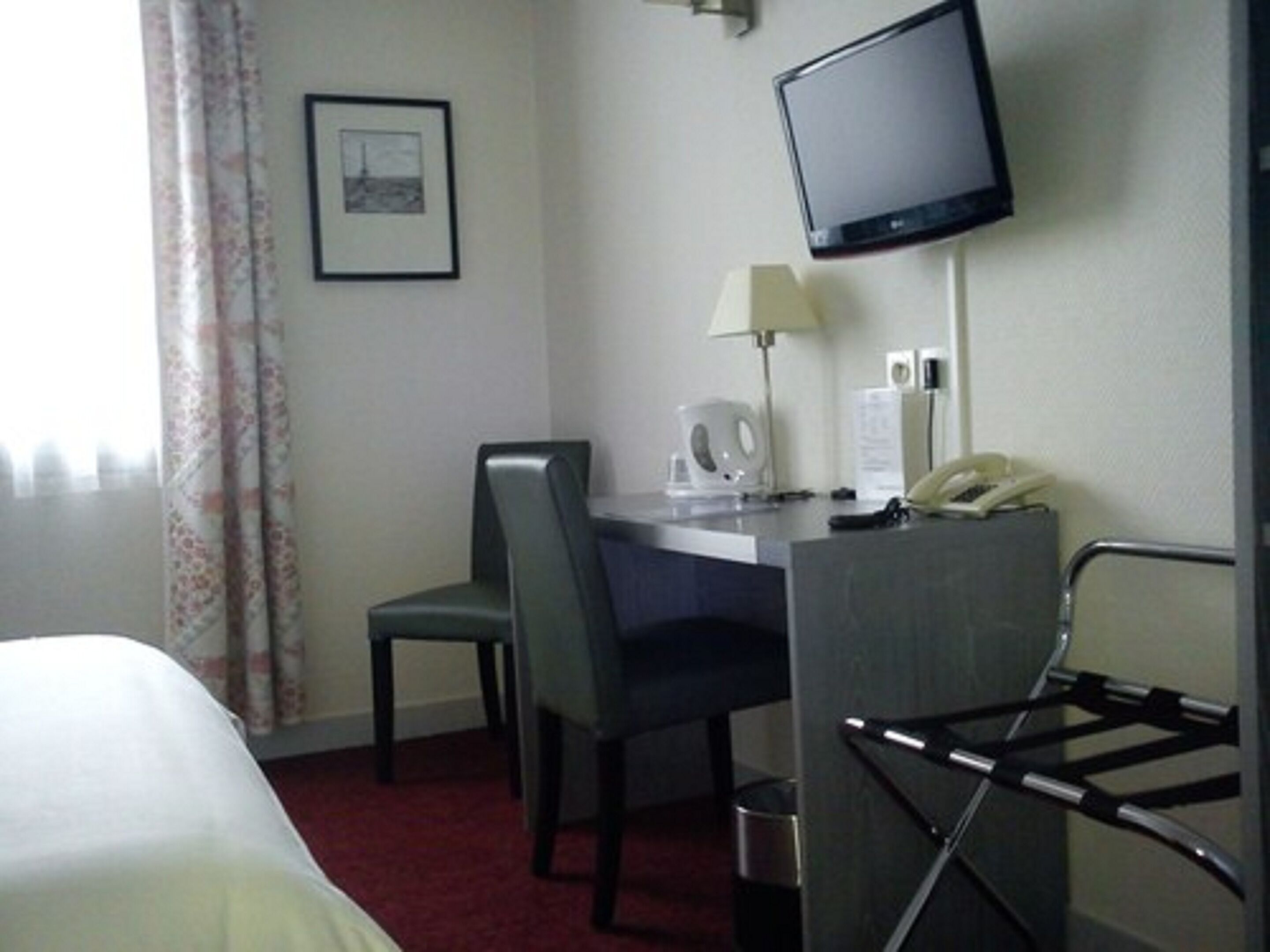 Una habitación de hotel con una cama, un escritorio con silla, un televisor montado en la pared, un teléfono y una ventana con cortinas.