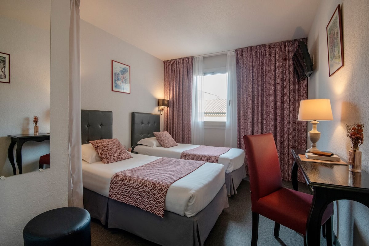 A hotel room with two beds, a desk, a chair, and a window with curtains.