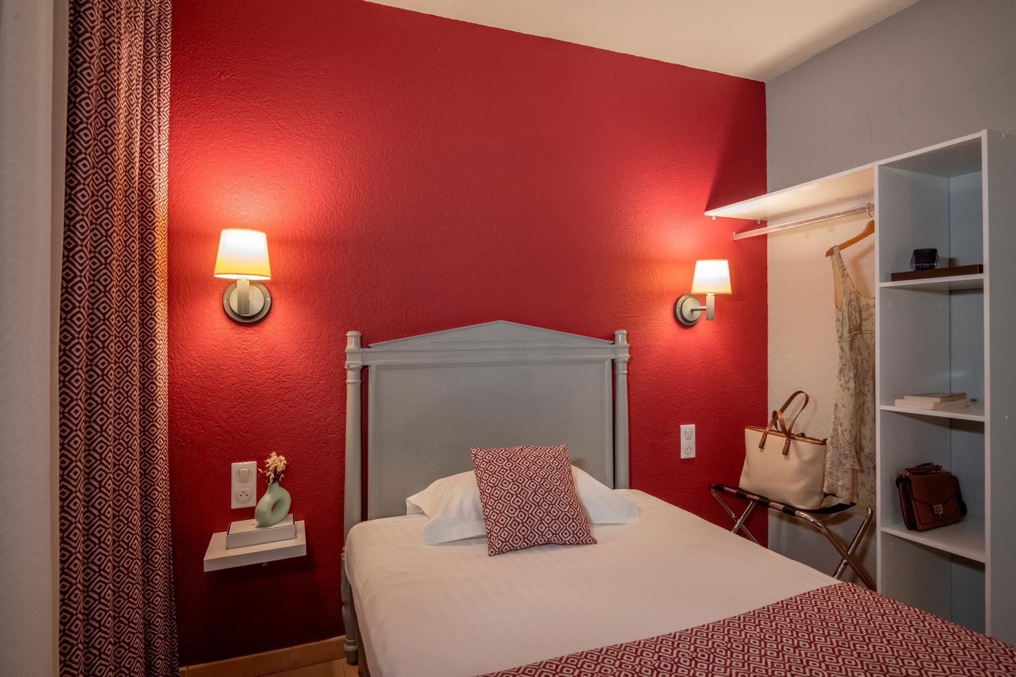 A hotel room with a red accent wall, a bed with white bedding, two bedside lamps, a nightstand with a vase, a wardrobe, and a hanging chair.