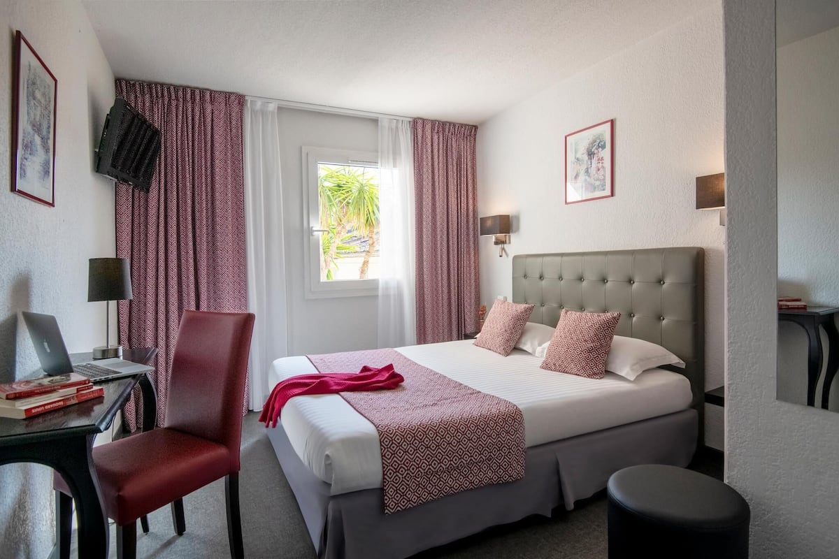 A hotel room with a bed, a desk with a laptop, a chair, and a small stool.