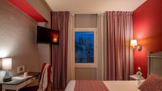 Une chambre d’hôtel comprenant un lit, un bureau, une chaise, une télévision, une fenêtre avec des rideaux, ainsi qu’une table de chevet avec une lampe et un vase.