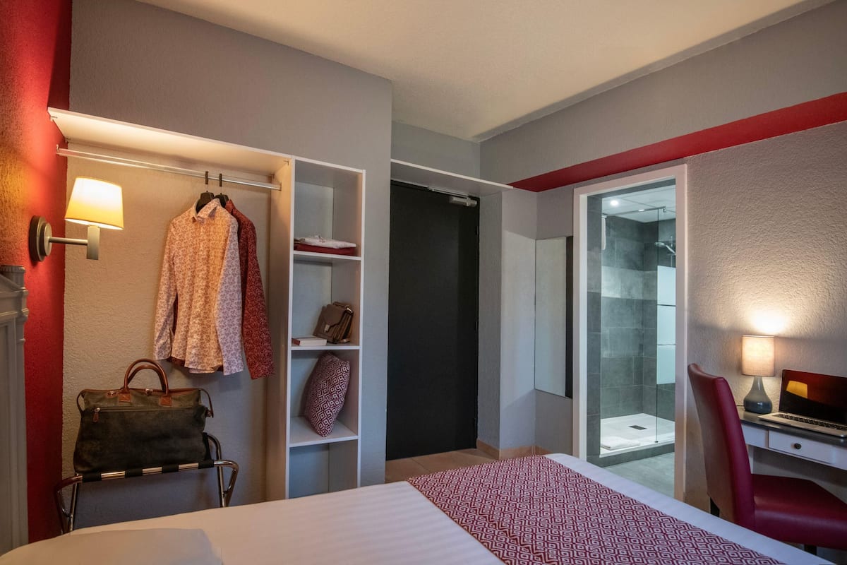A hotel room with a bed, a desk with a laptop, a chair, a wardrobe, a hanging coat rack, and a bathroom with a glass shower.