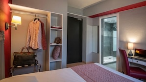 Ein Hotelzimmer mit Bett, Schreibtisch mit Laptop, Stuhl, Kleiderschrank und einer Garderobe sowie einem Badezimmer mit Duschkabine.