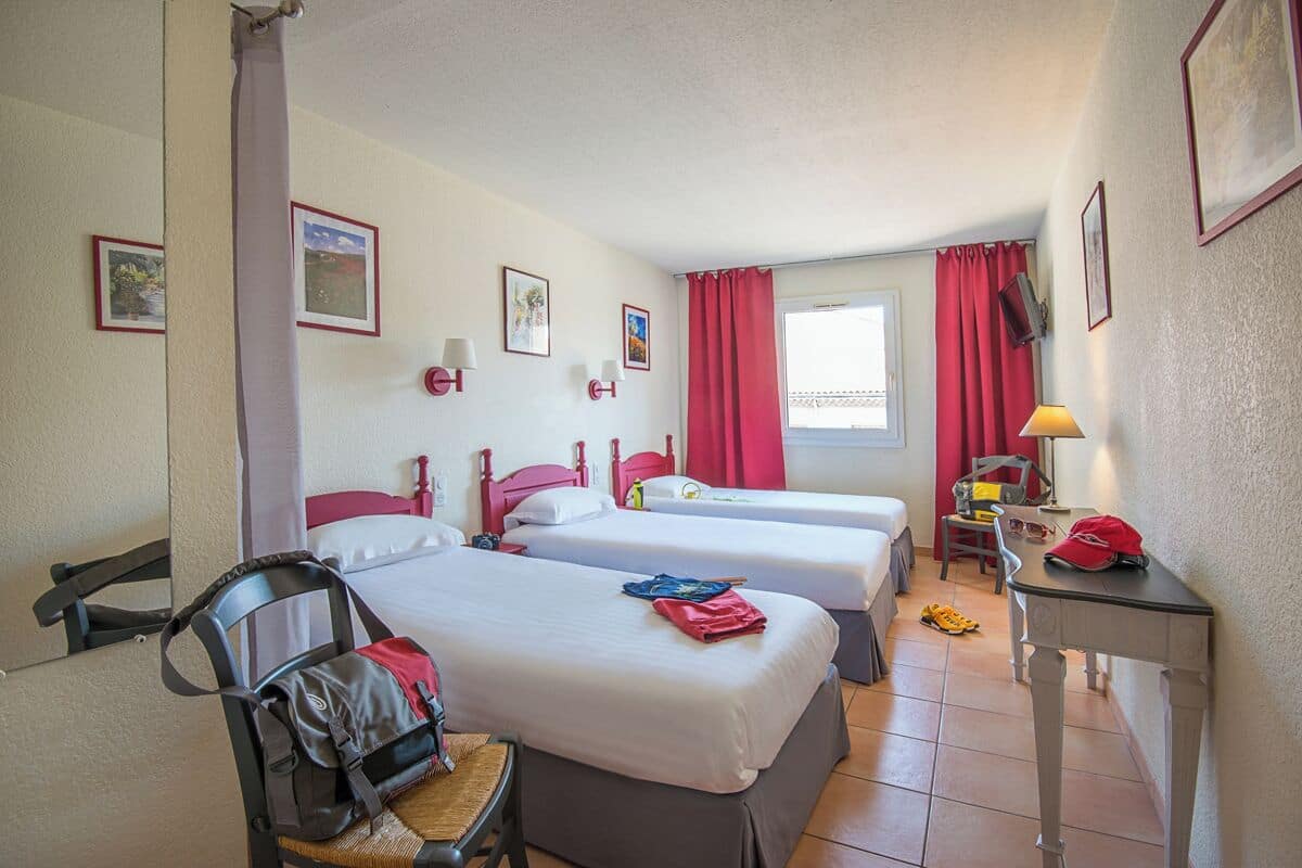 A hotel room with two single beds, a desk with a lamp, a chair, and a small table.