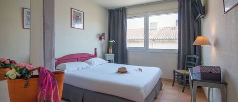 Un dormitorio con cama, una silla, una mesa, un ventanal con cortinas y un ramillete de flores.