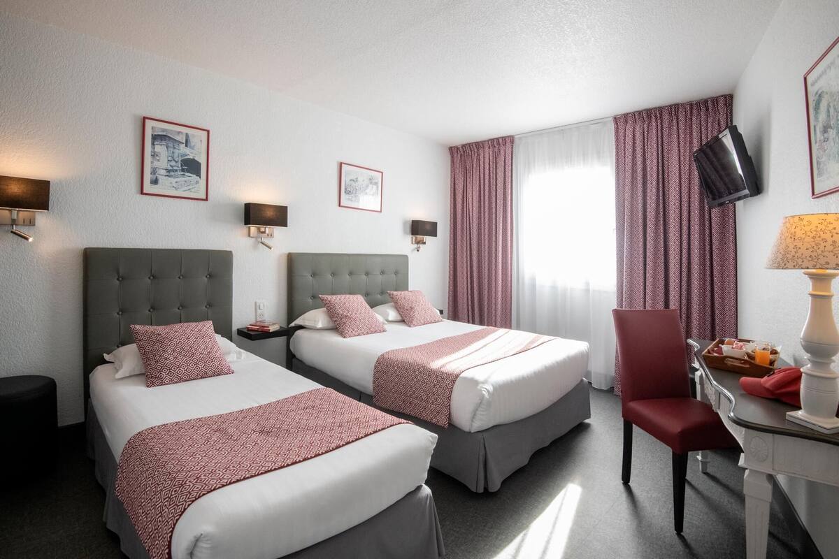 A hotel room with two beds, a desk with a lamp, a chair, and a TV mounted on the wall.