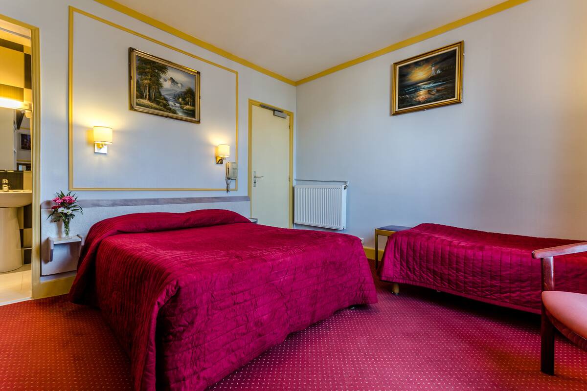 A hotel room with a large bed, a smaller bed, a desk, and a chair. There are two paintings on the walls and a radiator.