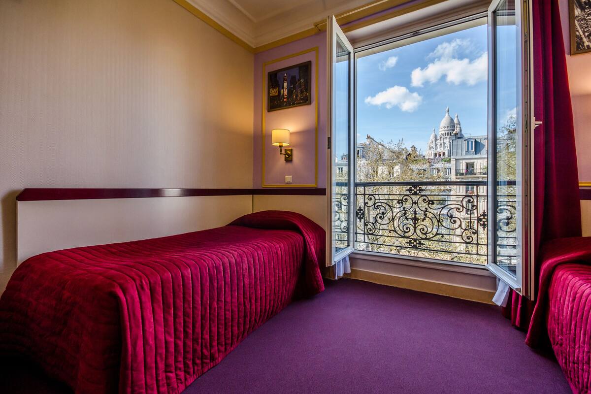 A hotel room with two beds, a large window with a view of a historic building, and a decorative wall lamp.