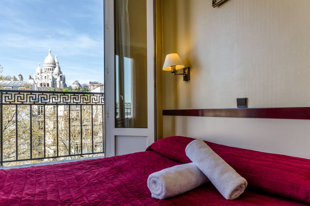A hotel room with a large window, a bed with a red quilt, a white towel, and a view of a domed building.