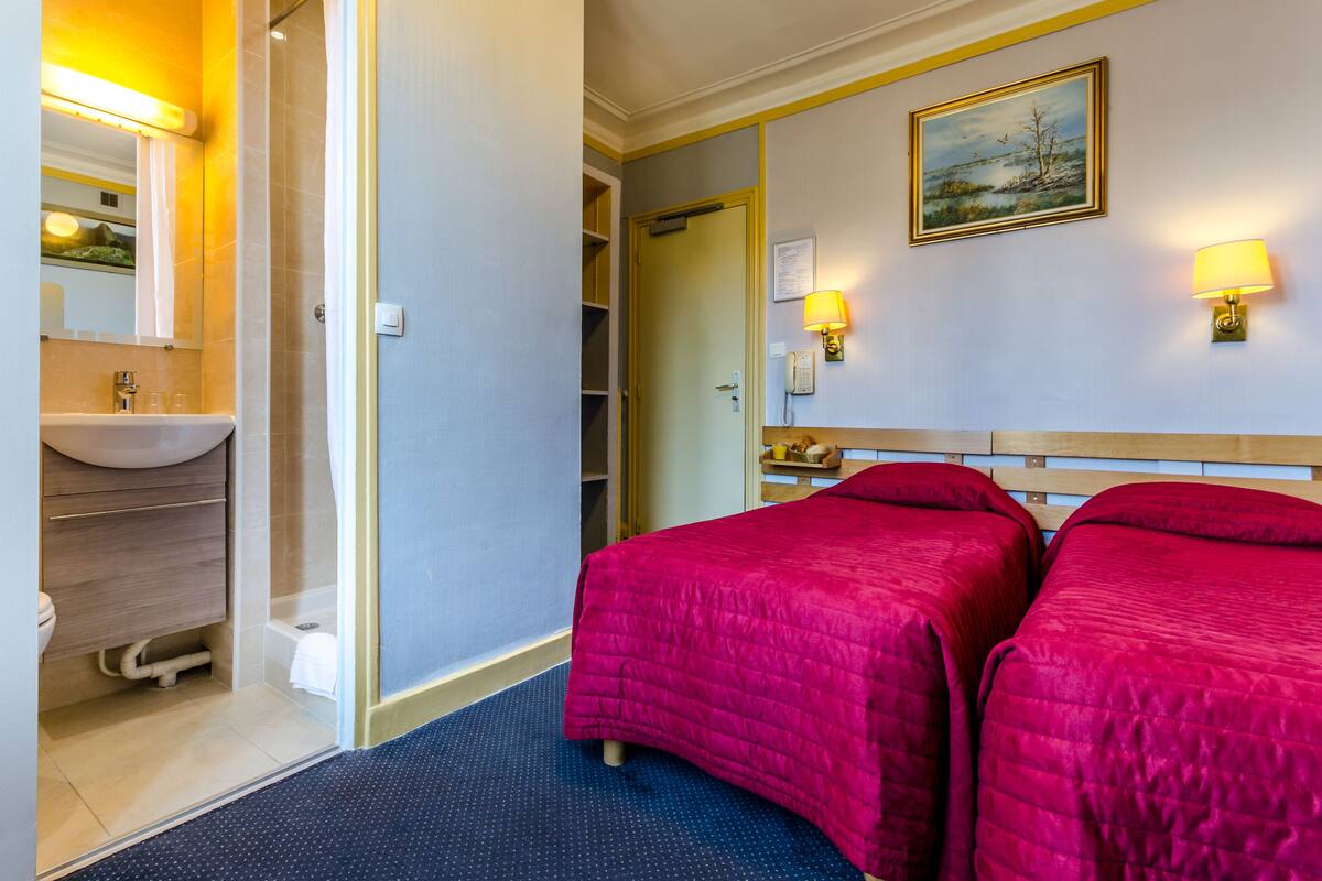 A hotel room with a bathroom, two beds with red bedding, a painting on the wall, and two wall-mounted lamps.