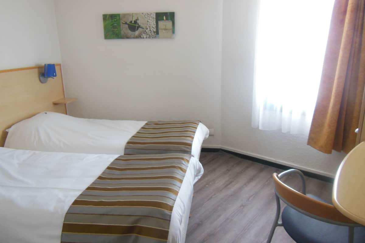 A hotel room with two beds, a small table, a chair, and a window with curtains.