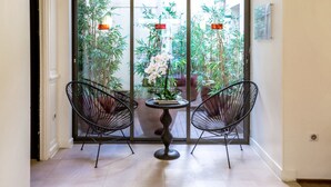 Una habitación con una puerta corrediza de vidrio que da a un jardín, dos sillas de alambre negras, una mesa redonda pequeña y una planta en maceta con flores blancas.