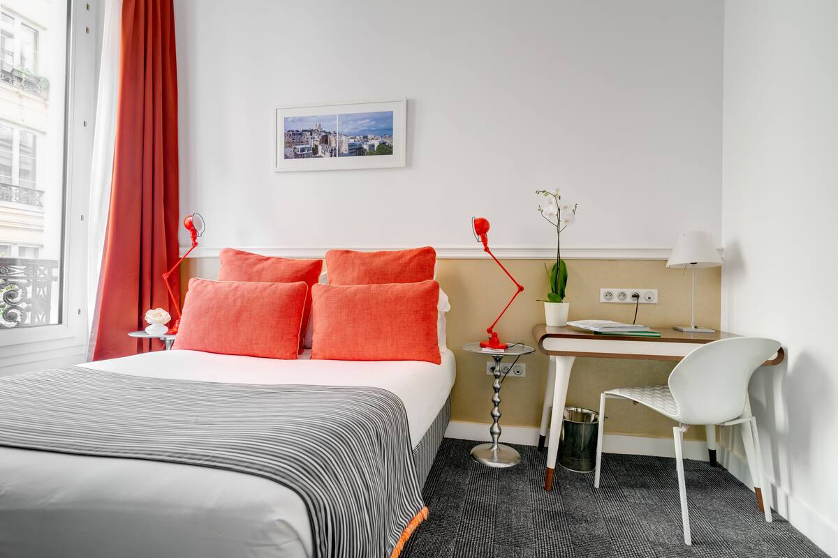Ein Hotelzimmer mit Bett, Schreibtisch, Stuhl, roter Lampe, kleinem Tisch mit Pflanze und Stadtbild an der Wand.
