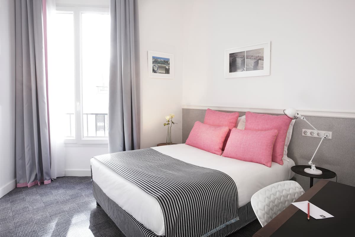 Ein Hotelzimmer mit einem Bett, einem Nachttisch, einem Schreibtisch, einer Lampe, einem Fenster mit Vorhängen und zwei gerahmten Bildern an der Wand.