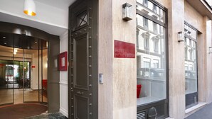 Entrada de un hotel con diseño moderno, que presenta un amplio escaparate de vidrio, un letrero rojo y una escalera de mármol.