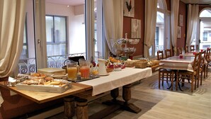 A breakfast buffet setup with various food items and beverages on a long table, surrounded by chairs and tables. - Logis Hotel Sully (Nogent-le-Rotrou)