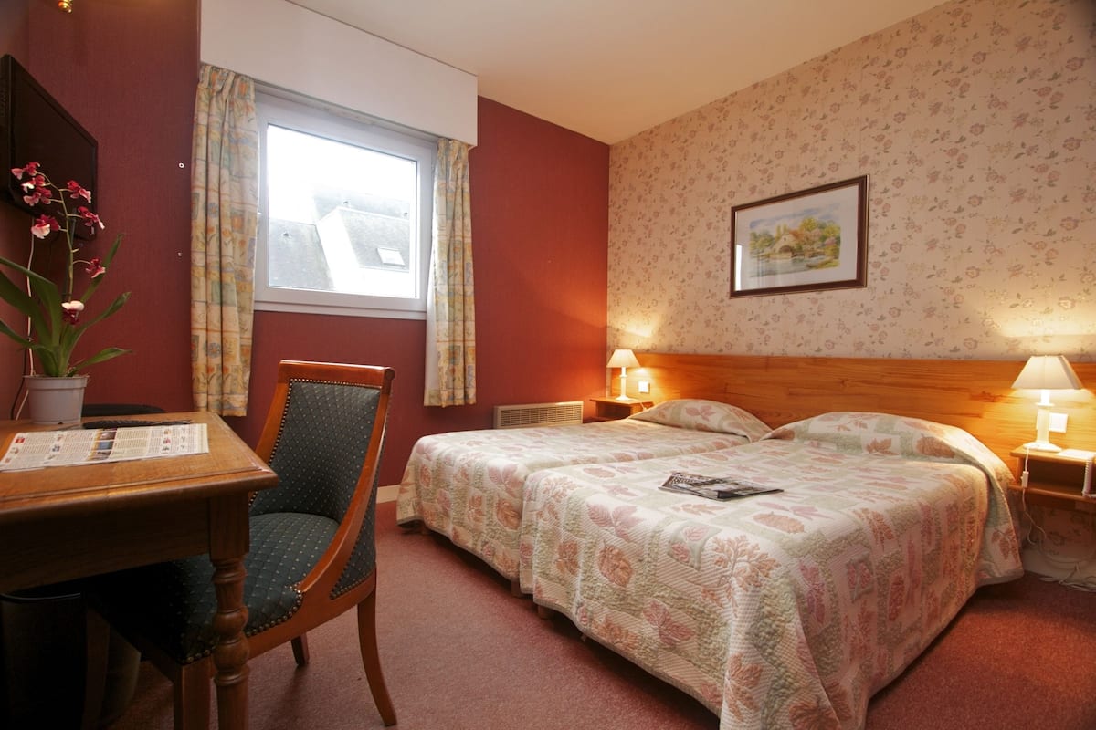 Una habitación de hotel con una cama, dos mesitas de noche con lámparas, una silla, un escritorio con una planta, una ventana con cortinas y un papel pintado con motivos florales.