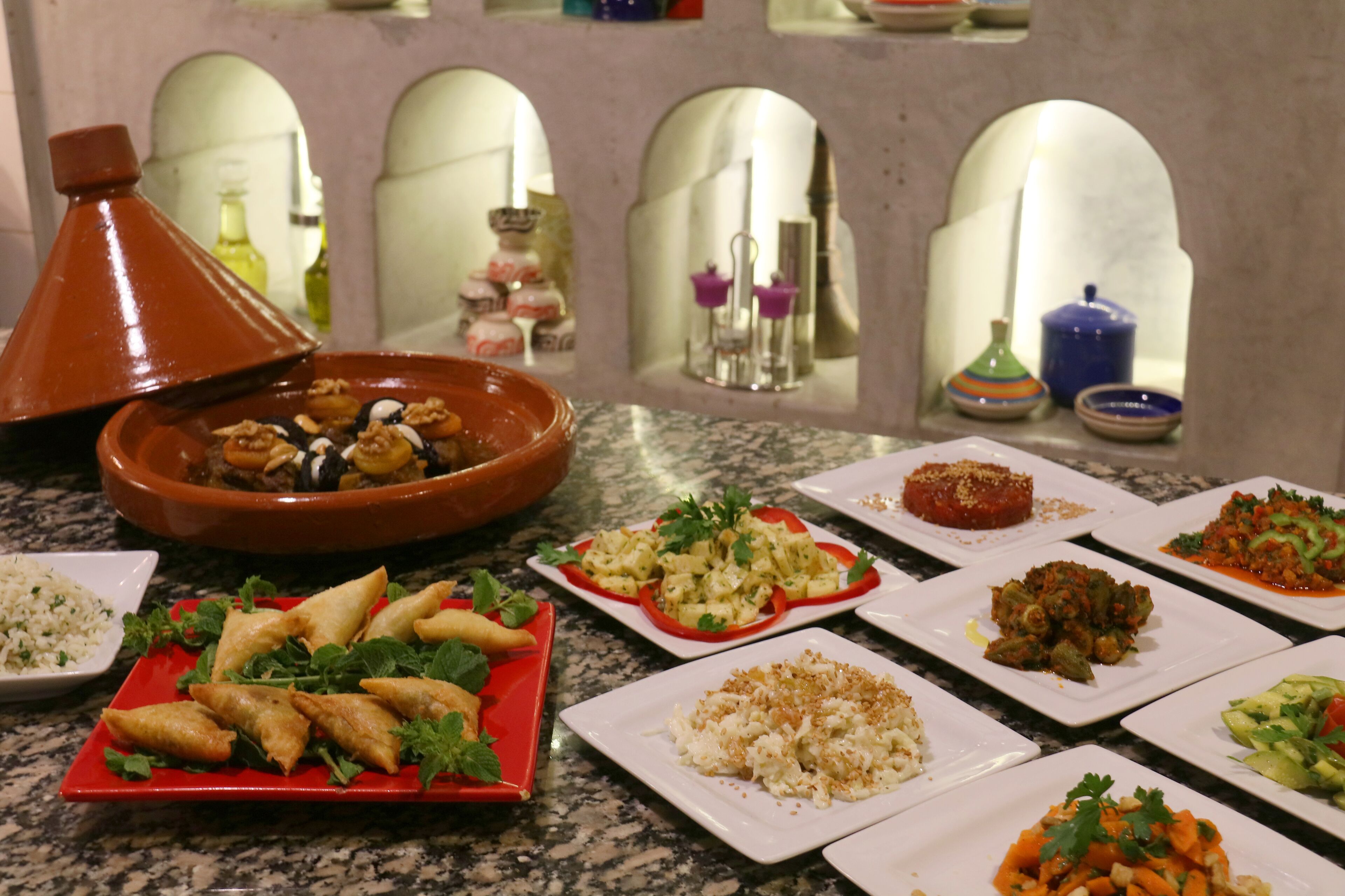 Diversi piatti su un tavolo in marmo, con una tajine marocchina e un arco decorativo sullo sfondo.