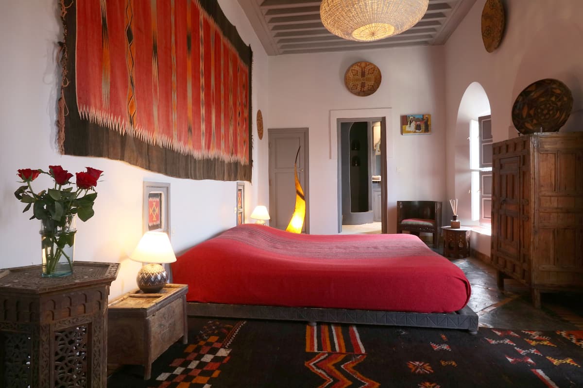 Ein Zimmer mit einem Bett, einer Lampe, einer Blumenvase, einem Teppich und einem Wandbehang.