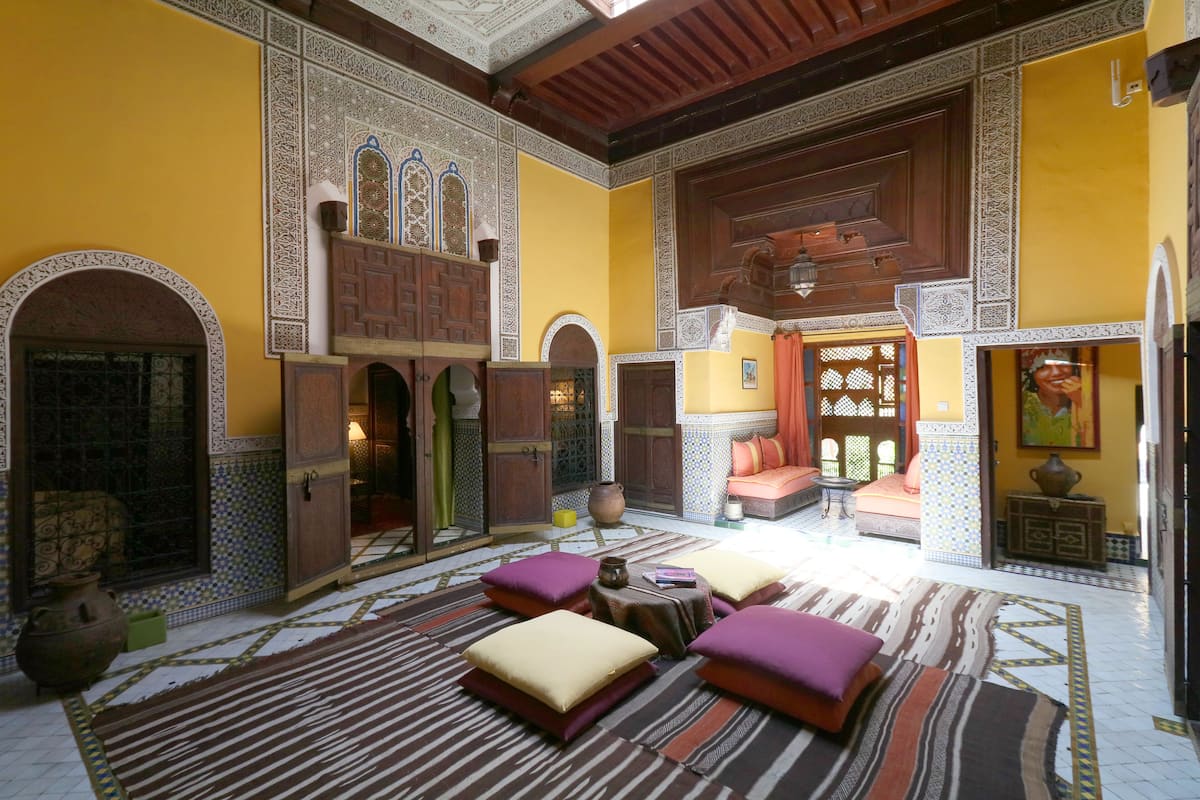 Ein traditionelles marokkanisches Interieur mit aufwendigen Fliesenarbeiten, Holzmöbeln und einer zentralen Sitzecke mit Kissen.