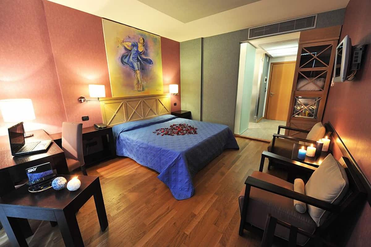 A hotel room with a bed, a desk with a laptop, a chair, a TV, and a painting on the wall.