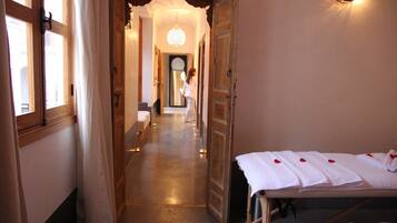 Un couloir d’hôtel avec des portes en bois, un passage menant à une chambre, et une table de massage avec des serviettes et des pétales de rose.