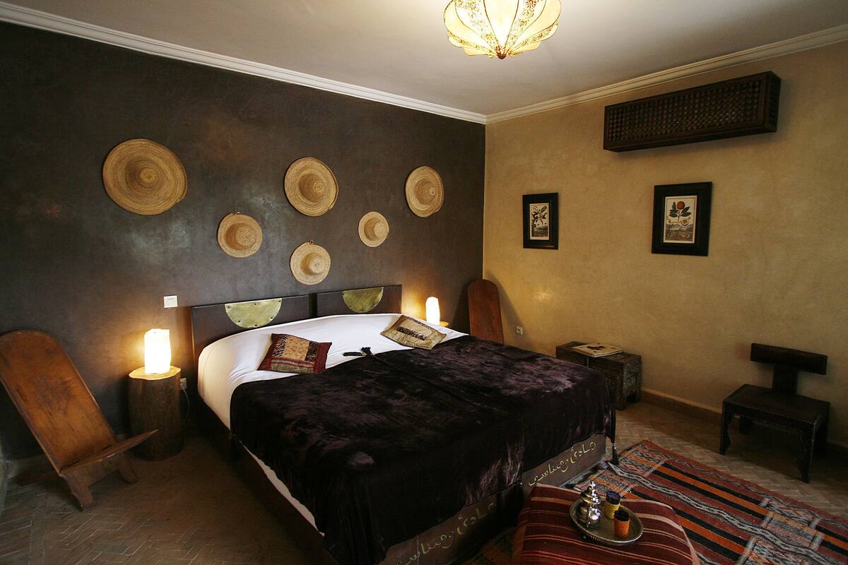 A bedroom with a bed, a wooden chair, a nightstand, a lamp, and a wall with decorative hats.