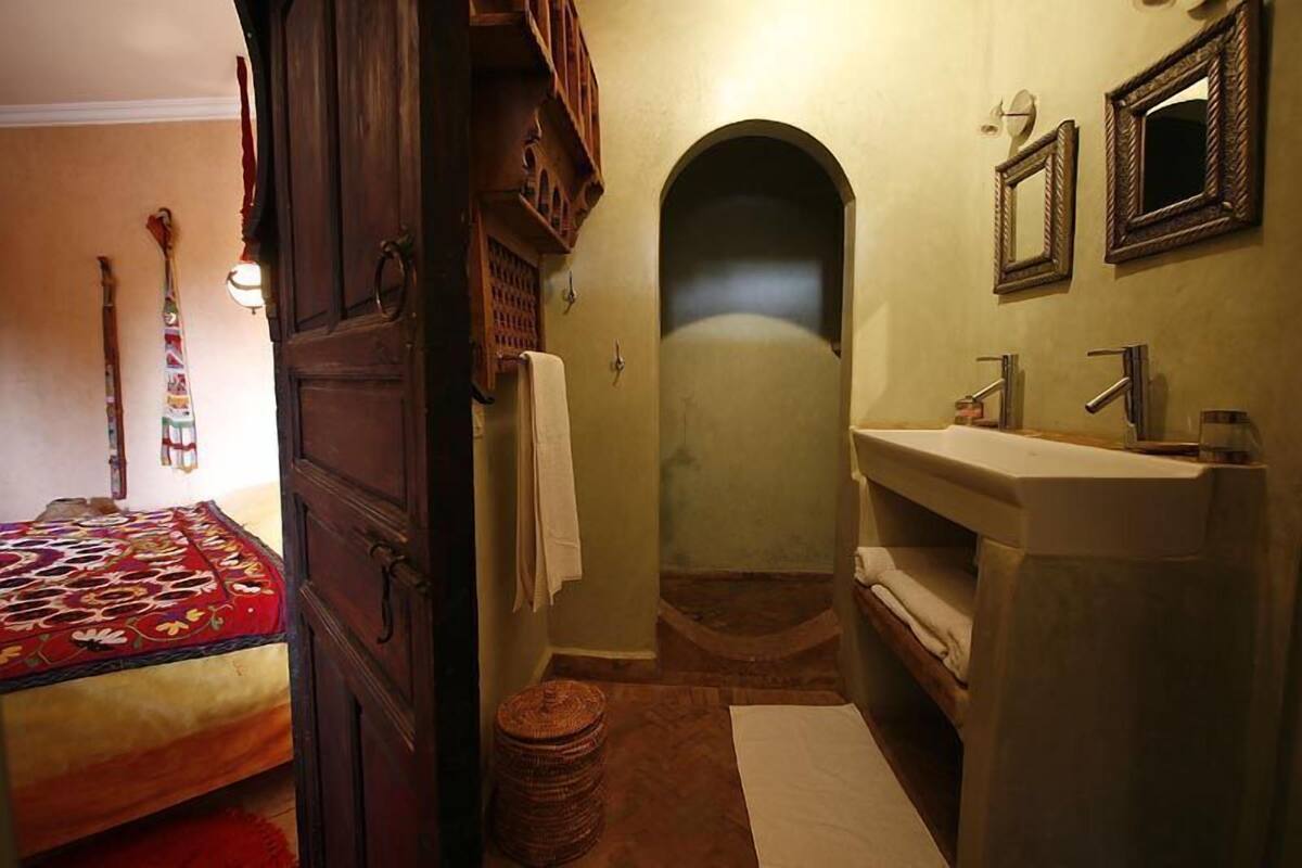 A narrow bathroom with a sink, mirror, and towel rack. A bed with a red blanket is visible in the adjacent room.
