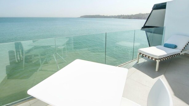 A balcony with a glass barrier overlooking a body of water, a white lounge chair, and a white table. - Hotel Lagon 2 (Dakar)