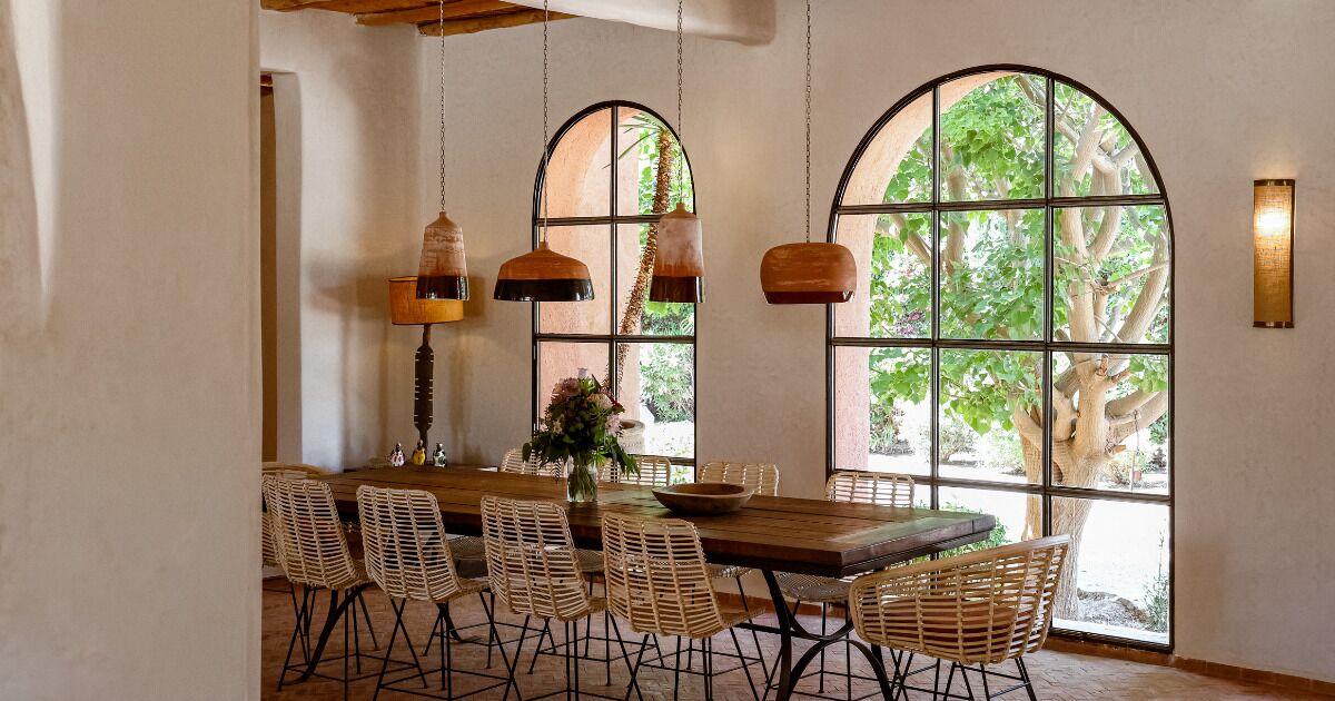 Zona pranzo con tavolo in legno, sedie in vimini, lampade a sospensione e ampie finestre ad arco che si affacciano sul verde circostante.
