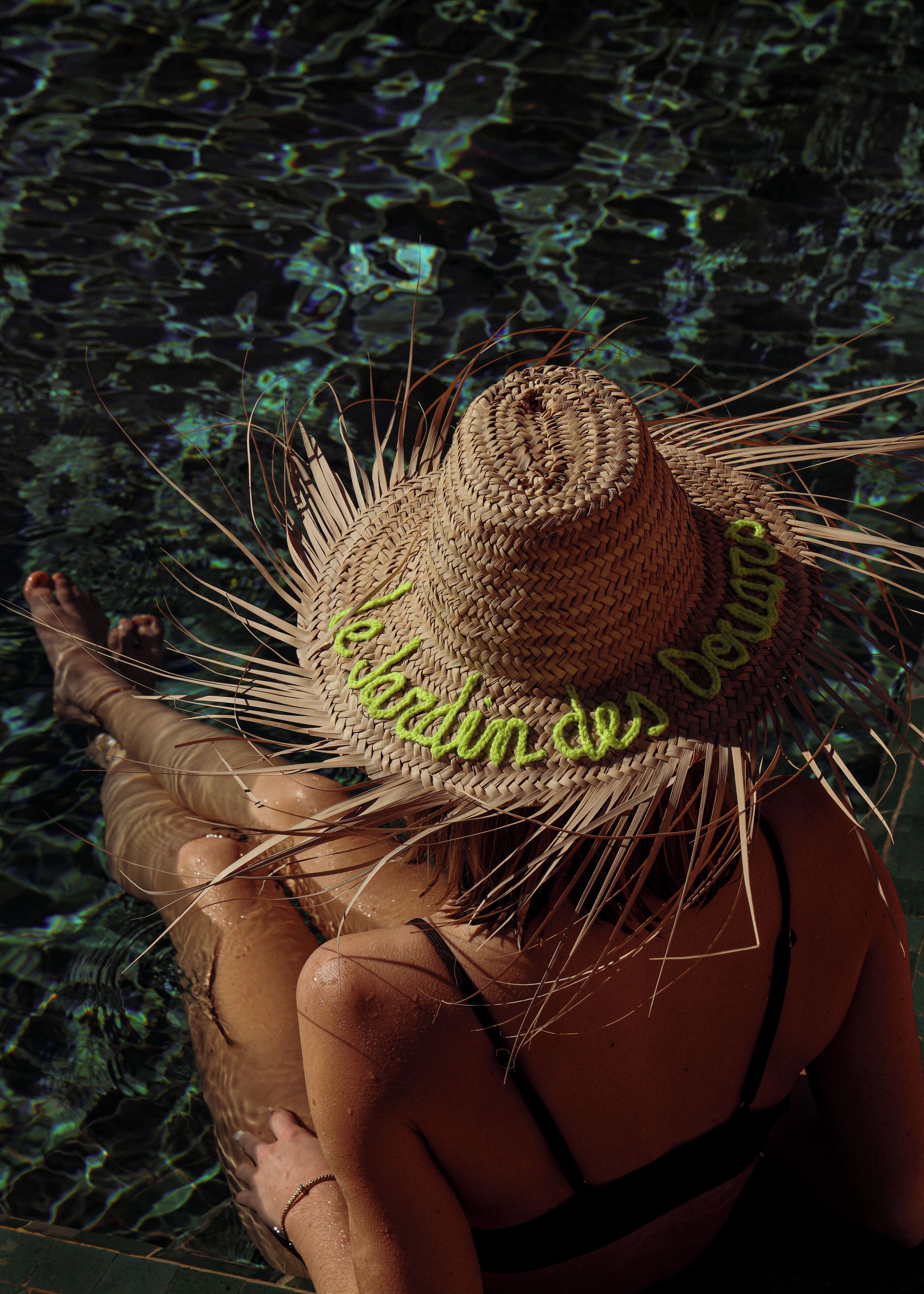 A person wearing a straw hat with a woven pattern and a green inscription, standing by a body of water.